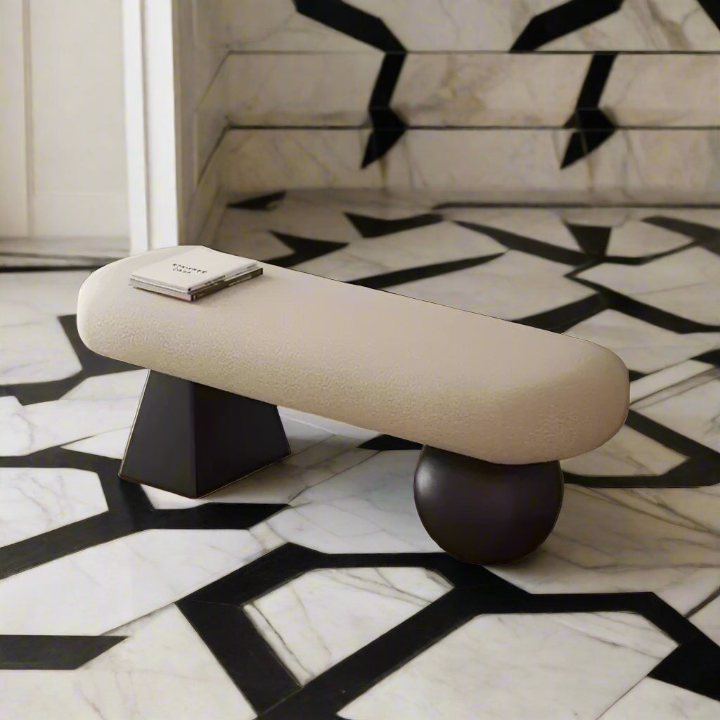 Beige bench with black spherical base in a room with wooden floor and framed pictures on the wall.