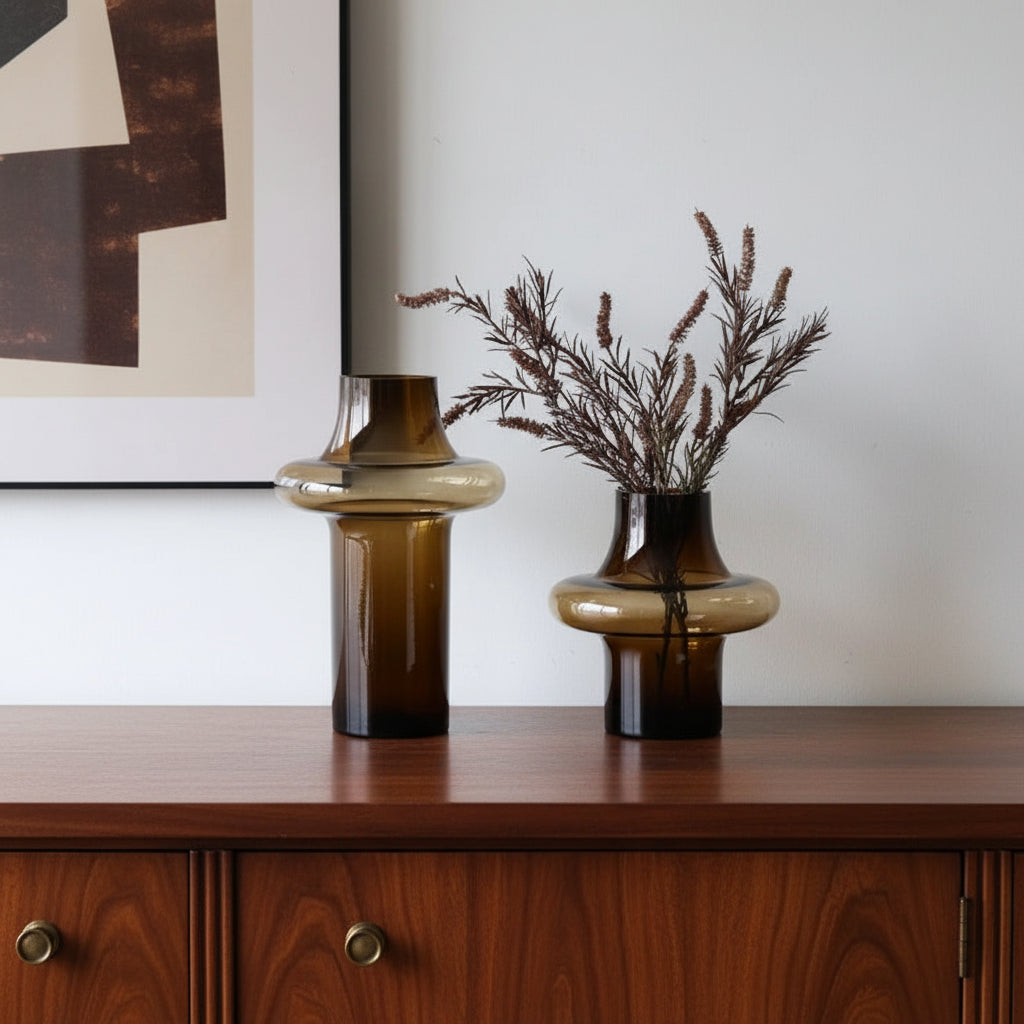 Two brown glass vases with one containing dried plants on a white surface.