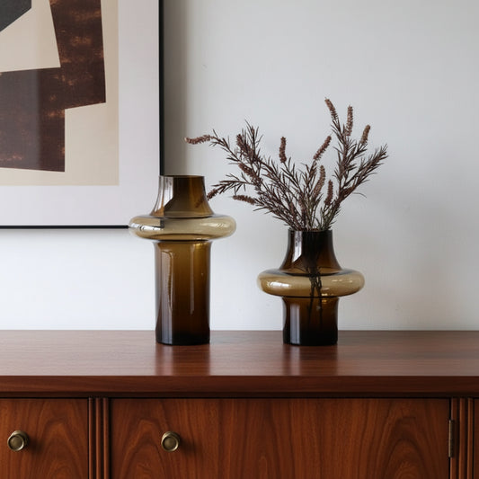 Two brown glass vases with one containing dried plants on a white surface.